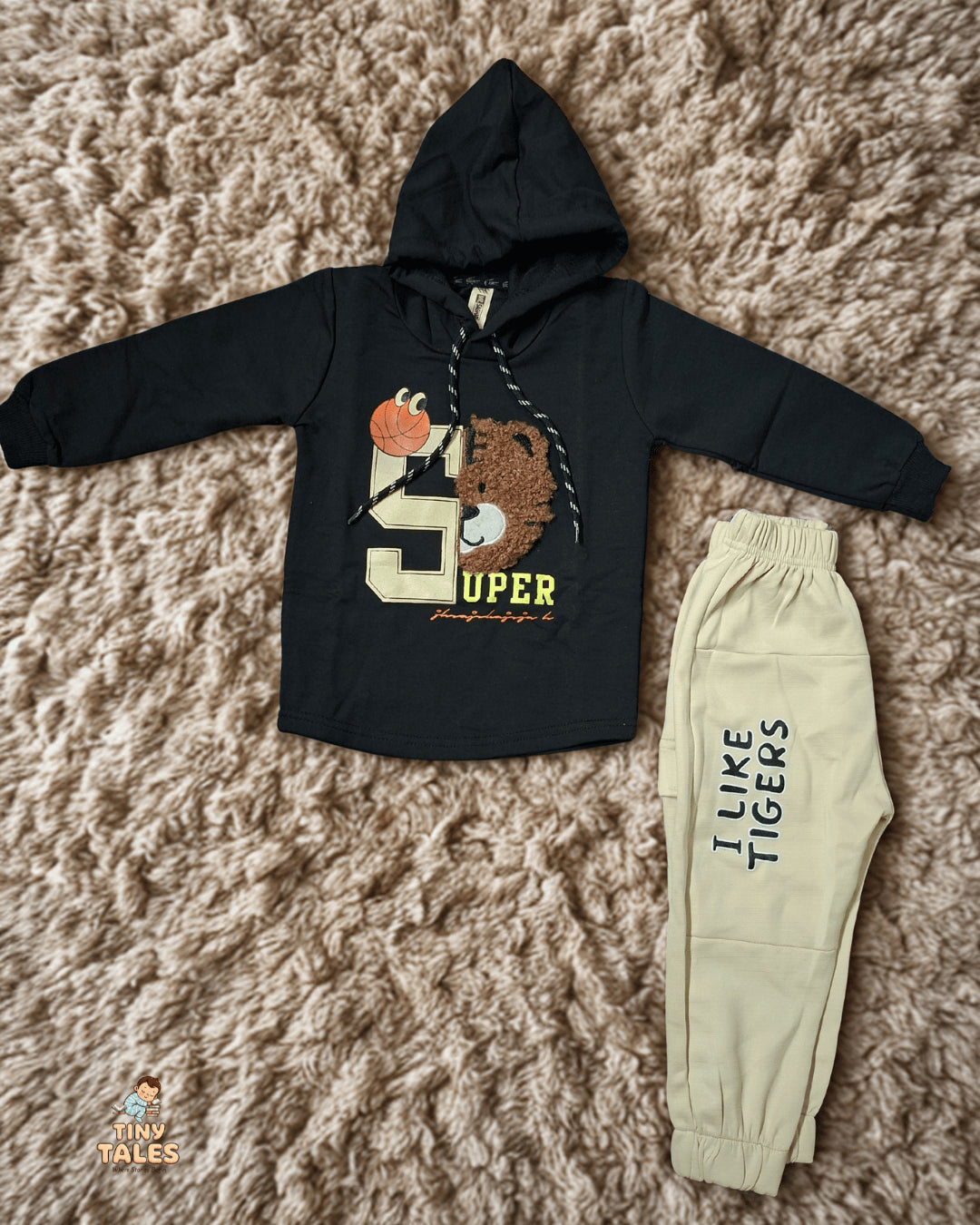 Black hoodie with a tiger design and beige pants on a textured surface. Tiny Tales baby clothing, Organic baby clothes Pakistan, Premium baby wear, Soft cotton baby rompers, Newborn and toddler outfits, Baby dresses and essentials, Eco-friendly baby fashion, Cute baby clothes online, Safe baby clothing brand, Tiny Tales baby shop